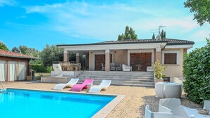 Un espace aménagé au bord de la piscine, avec des chaises longues, un patio couvert et un chemin en briques menant à un bâtiment doté d’un toit en tuiles.