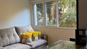 A modern living room with a grey sofa, yellow cushions, a glass coffee table, and a large window with a view of greenery. - Comfortable apartment with a view of nature (Porto Alegre)