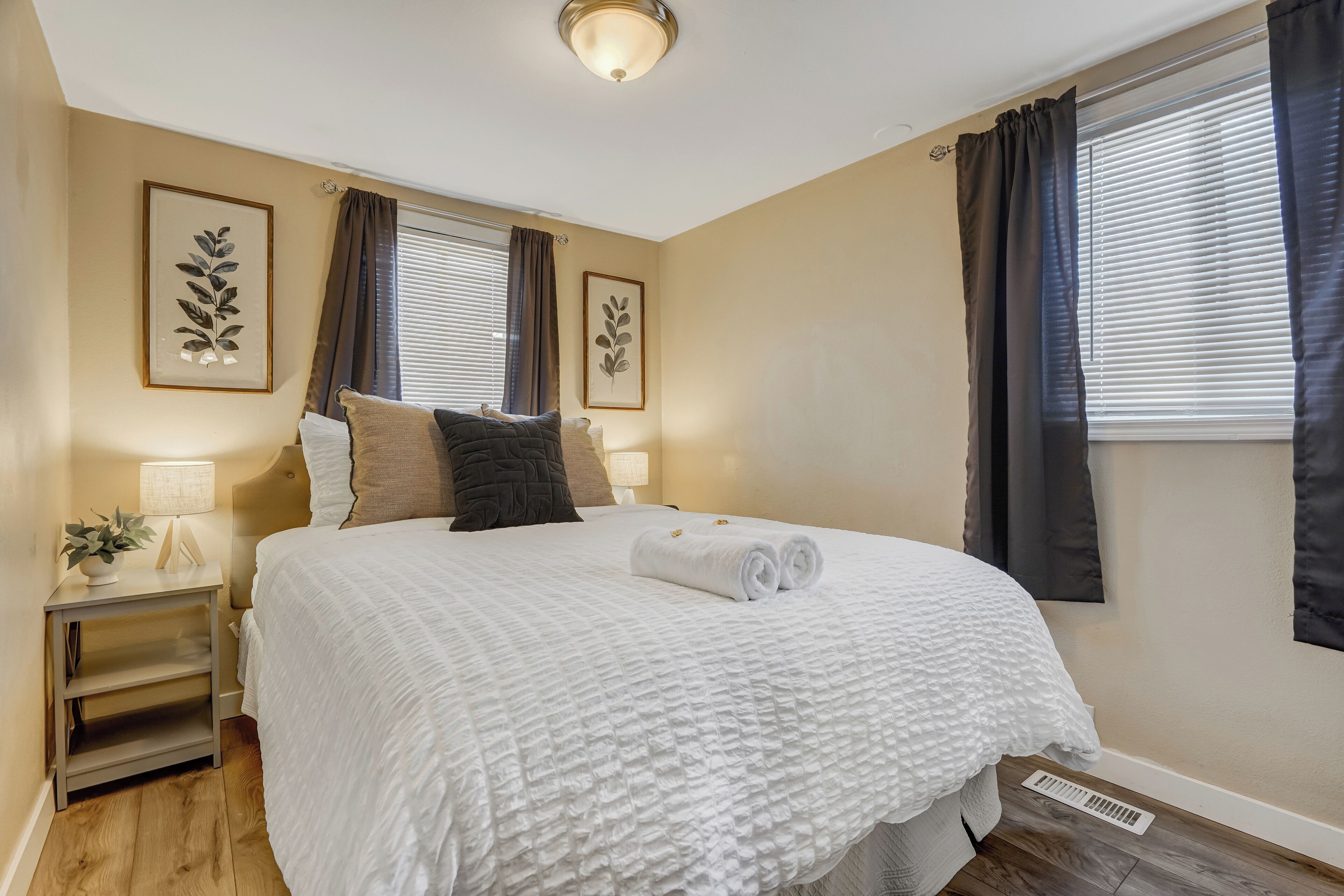 A neatly made bed with a white quilt, two pillows, and a rolled towel. There are two framed leaf artworks on the wall, a bedside table with a lamp, and a window with curtains.