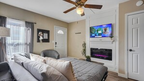 A bedroom with a bed, a chair, a TV mounted above a fireplace, and a ceiling fan. - Downtown Urban Basecamp - Private Patio, Grill and Fireplace (Colorado Springs)