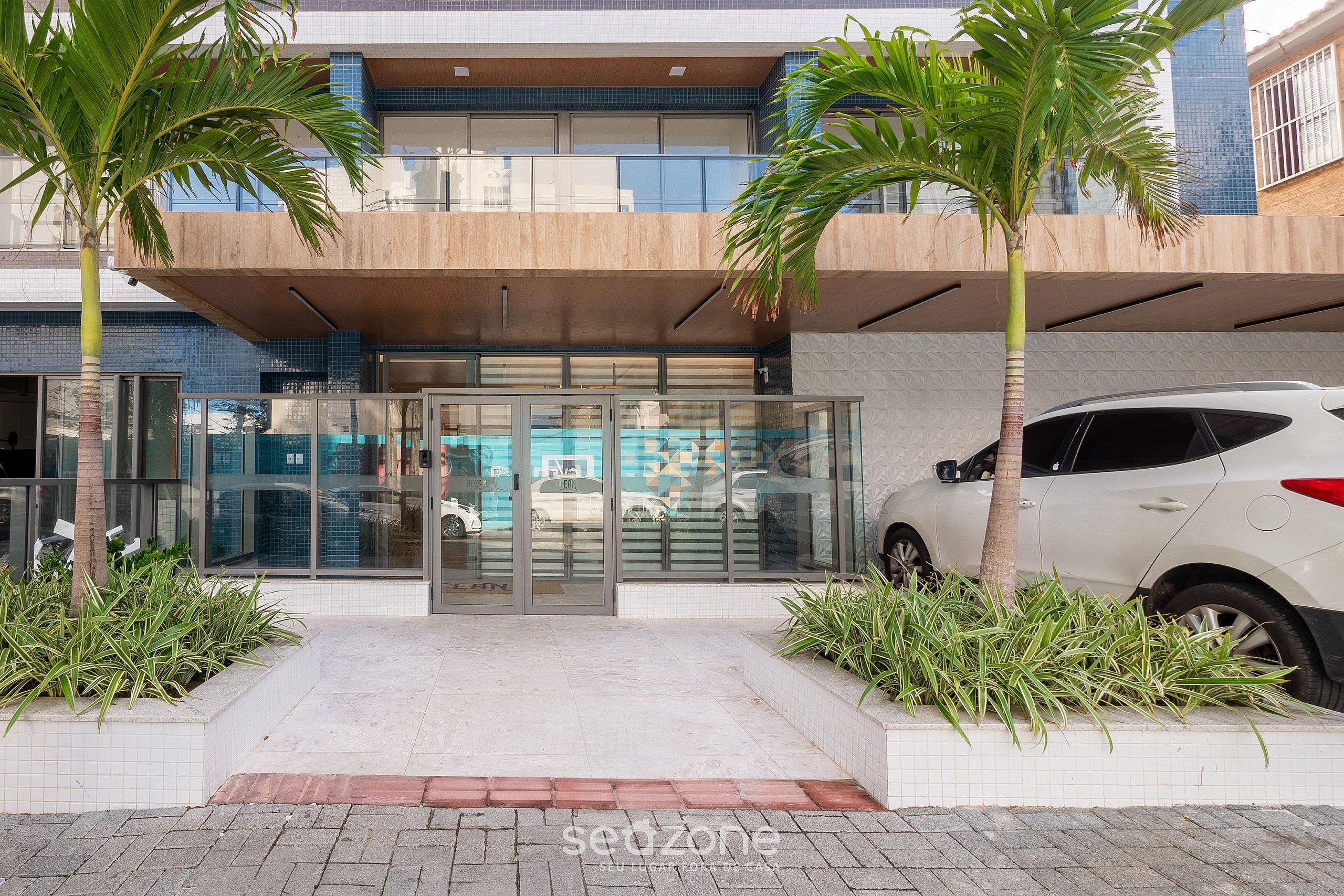 A modern building with a glass entrance, a white car parked outside, and palm trees.
