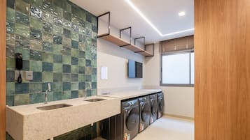 A modern bathroom with a tiled backsplash, a dual sink vanity, a mirror, a wall-mounted television, and a window with blinds.