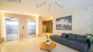 A modern lobby with two elevators, a grey sofa, a coffee table with a flower, and a large wave painting on the wall.