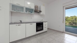 A modern kitchen with white cabinets, a built-in oven, and a view of a green landscape through a sliding door. - Cozy apartment in Krusevo with WiFi (Krusevo)