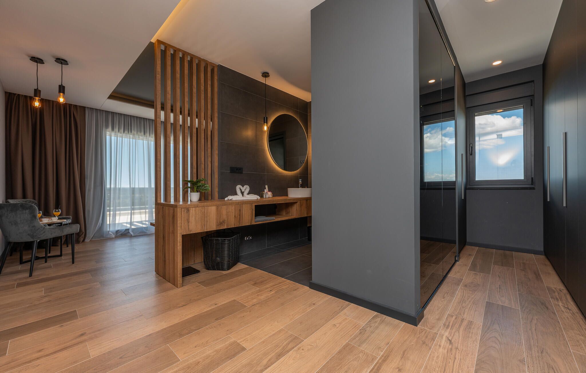 A modern bathroom with a wooden vanity, a round mirror, and a large window.