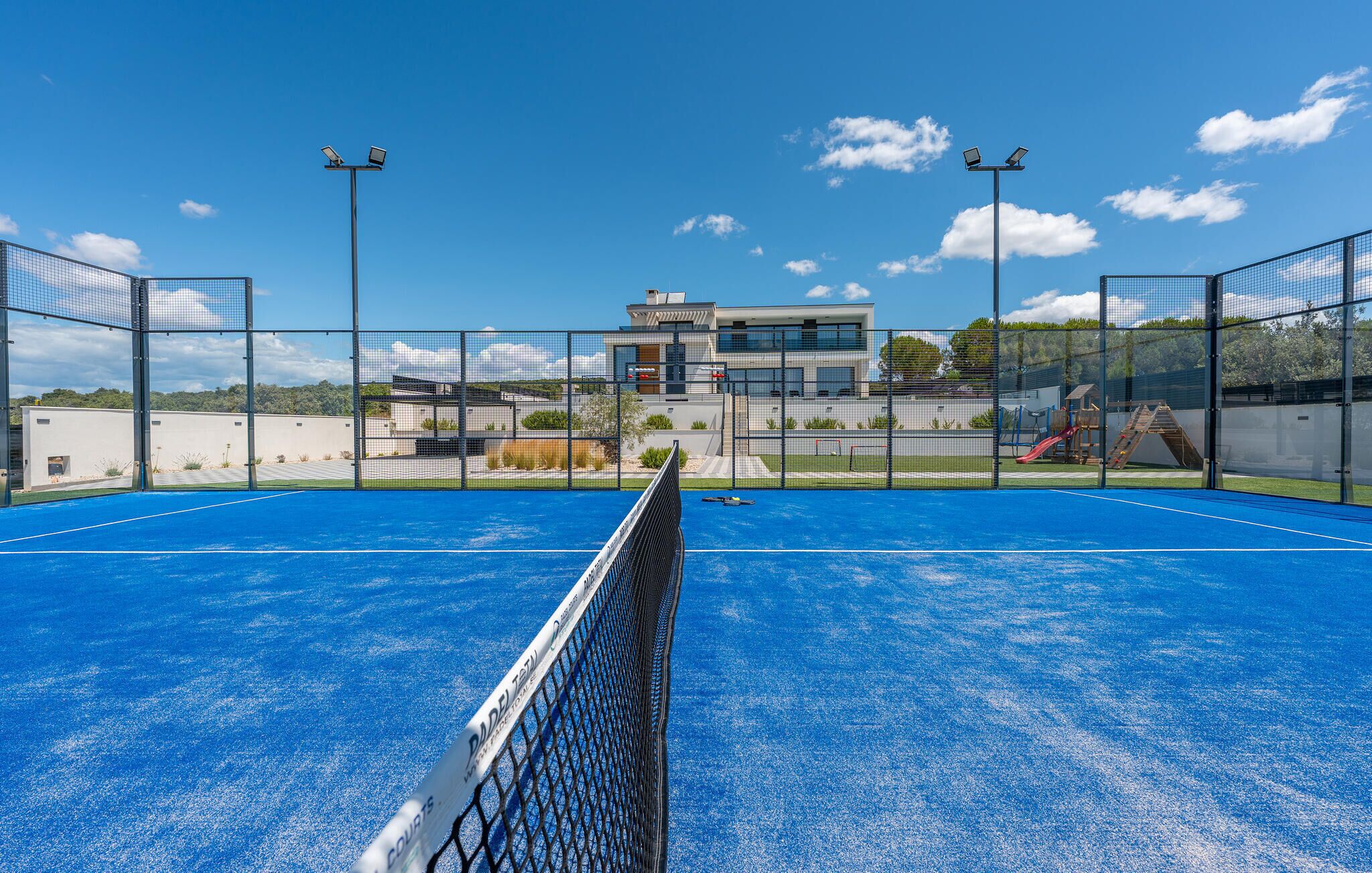 An outdoor tennis court with a net in the foreground, surrounded by a fence, and a modern building in the background.