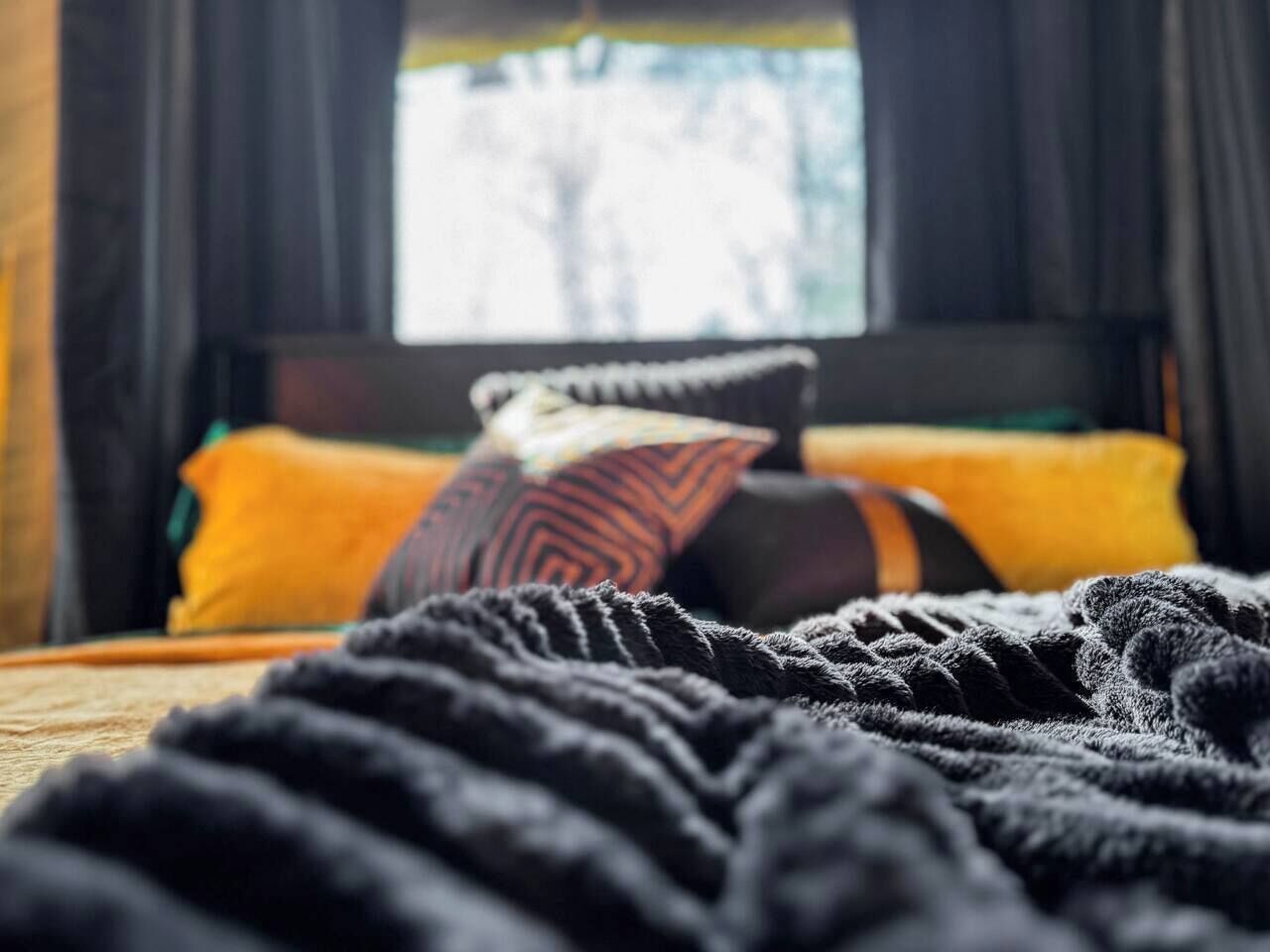 Un lit avec une couverture et des oreillers sombres, une fenêtre avec des rideaux et une vue sur les arbres à l’extérieur.