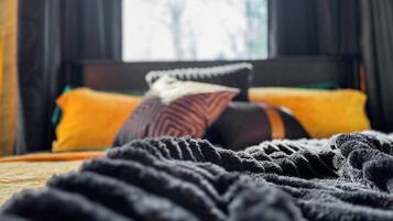 Un lit avec une couverture et des oreillers sombres, une fenêtre avec des rideaux et une vue sur les arbres à l’extérieur.