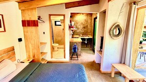 A bedroom with a large bed, wooden ceiling beams, a small bench, and a view into a kitchen area.