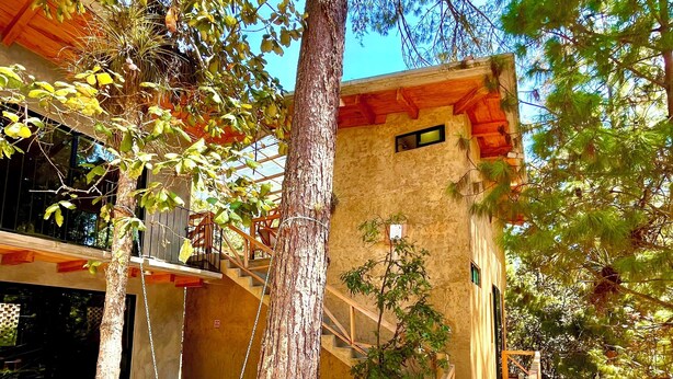 A rustic cabin with a wooden staircase leading to a balcony, surrounded by trees and a gravel ground.
