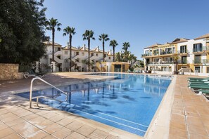 A swimming pool with a metal ladder, surrounded by palm trees and a building with balconies.