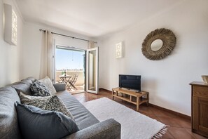 Ein modernes Wohnzimmer mit einem grauen Sofa, einem weißen Teppich, einem Fernseher auf einem Holzboden und einem Balkon mit Tisch und Stühlen.
