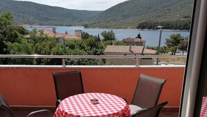 A balcony with a table and chairs, overlooking a lake with buildings and hills in the background.