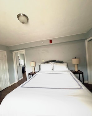 Une chambre d’hôtel avec un grand lit, deux tables de chevet avec des lampes, un luminaire au plafond et une porte donnant sur une autre pièce.