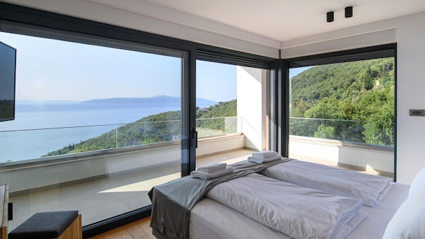 A modern bedroom with a large bed, a small seating area, and a balcony with a scenic view of the sea and hills.