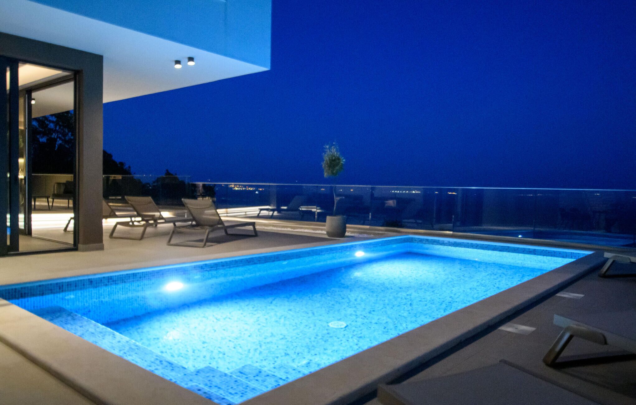 Une piscine dotée de chaises longues et d’une balustrade en verre, entourée d’un bâtiment moderne offrant une vue sur le ciel nocturne.