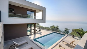A modern two-story house with a rooftop infinity pool, outdoor seating, and a view of the sea.
