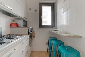 A narrow kitchen with white cabinets, a microwave, a gas stove, and teal stools.