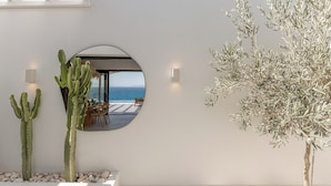 Een modern interieur met een grote ronde spiegel die een eethoek en uitzicht op zee weerspiegelt. Er staan twee cactussen en een olijfboom.