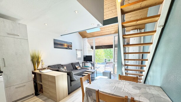 A modern kitchen with a built-in oven, a dining area with a wooden table and chairs, a grey sofa, and a view of the outdoors.