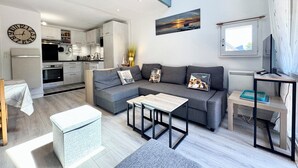 A modern living area with a grey sofa, a coffee table, and a TV stand. A kitchen is visible in the background.