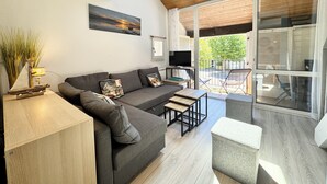 A modern living room with a grey sofa, a small wooden table, and a TV. There is a painting on the wall and a window above the sofa.