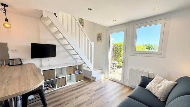 A modern living room with a staircase, a flat-screen TV, a dining table, and a sofa with cushions. - Maison refaite à neuf - Piscine et parking privé (Guidel)