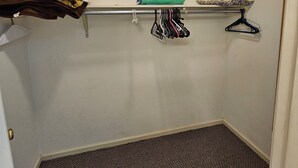 Een kleine, zwak verlichte kamer met een kamerbreed tapijt, een metalen plank met opgevouwen kleding en een deur met een messing handgreep.