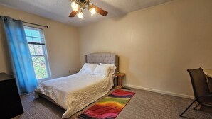 Een slaapkamer met een bed, een plafondventilator, een raam met gordijnen, een nachtkastje en een stoel.