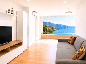 A modern living room with a flat-screen TV, a grey sofa with patterned cushions, and a large window offering a coastal view. - 🏖Sea views + private beach. What else? (Tossa de Mar)