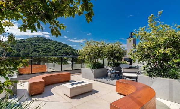 A rooftop terrace with curved seating, a coffee table, and a view of a forested hill. - Building With Swimming Pool Next to Balneario Shopping in BC - TSO (Balneário Camboriú)