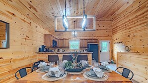 Interior de una cabaña de madera con una mesa de comedor para cuatro, una zona de cocina y un ventanal grande.