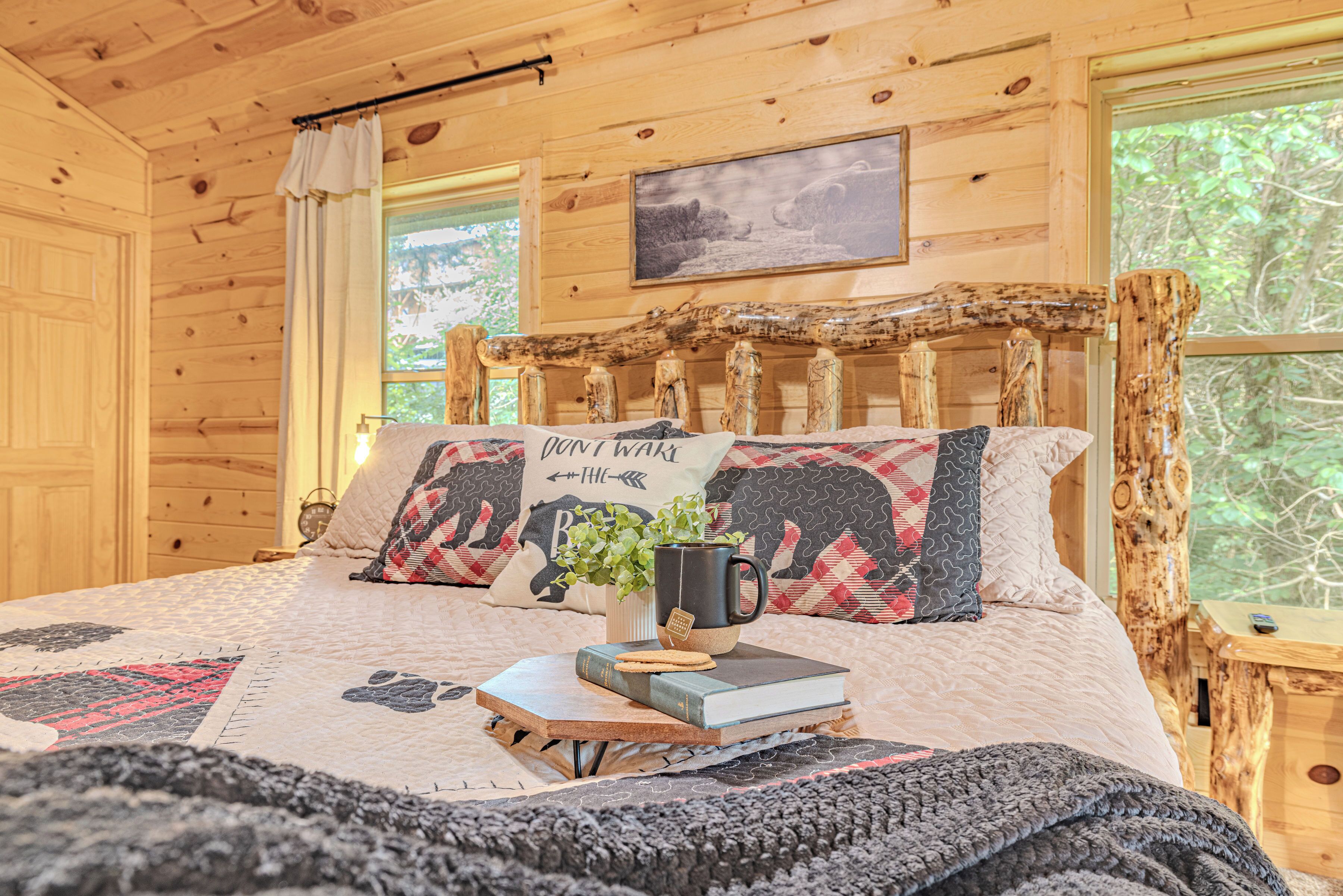 A cozy wooden cabin bedroom with a large bed, a quilted blanket, a pillow with a bear design, a mug, and a book.