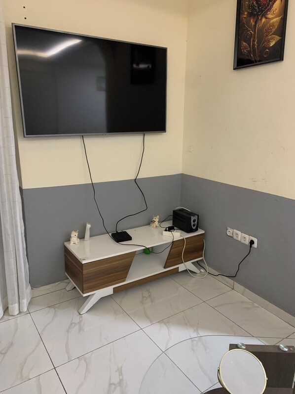 A room with a flat-screen TV mounted on the wall, a wooden and white desk with a small figurine, a radio, and a power strip.