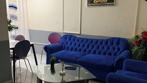A living room with a blue sofa, a glass coffee table, and a chair. There is a painting on the wall and a potted plant on the table. - DCHhome available for your best stays in the city of Douala (Douala)