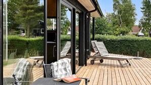 A covered outdoor deck with lounge chairs and a table, surrounded by greenery.