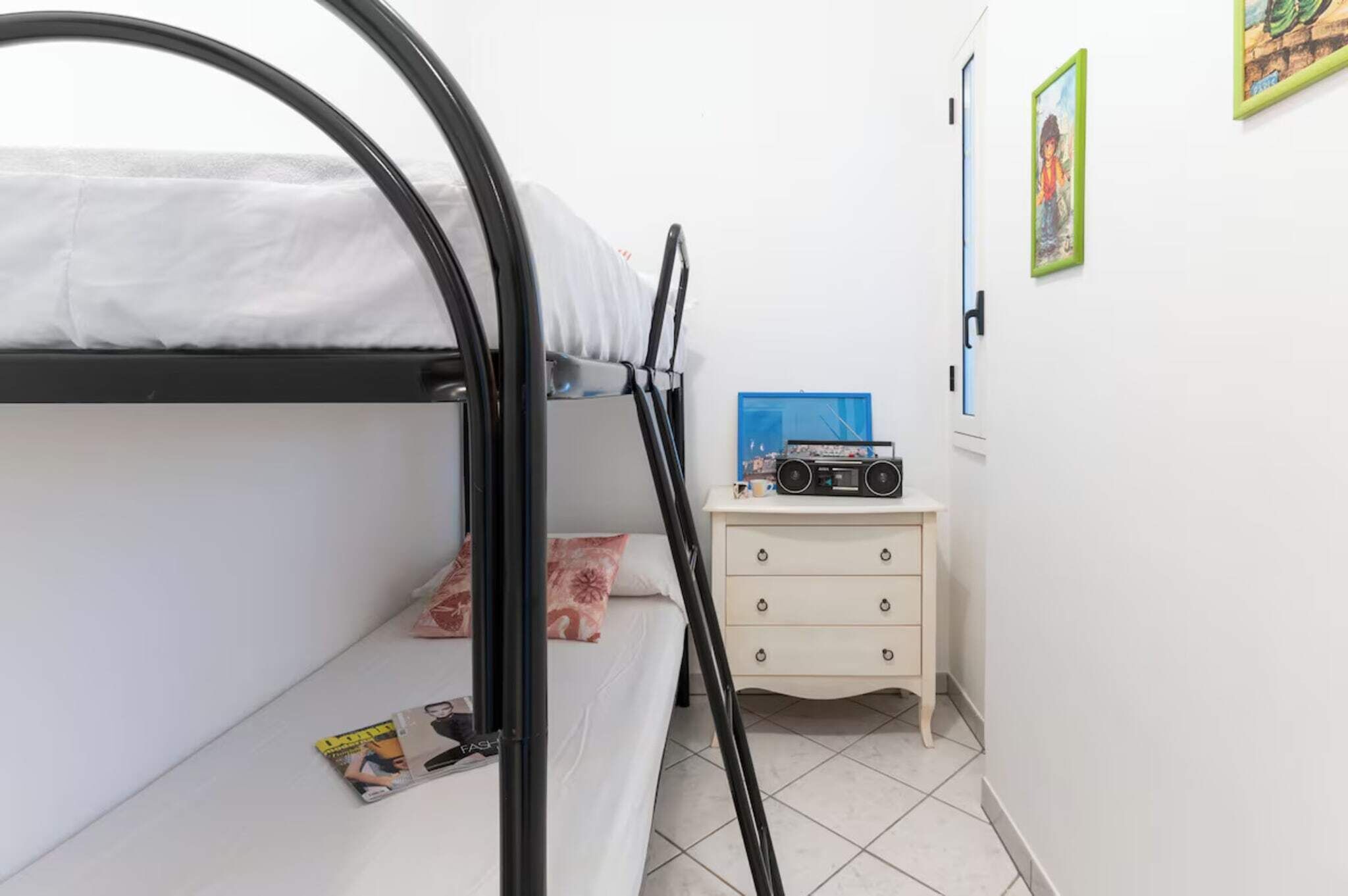 Ein Etagenbett mit einer Zeitschrift und einem Kopfkissen auf der unteren Ebene, eine Kommode mit einem Radio und ein Bilderrahmen an der Wand.