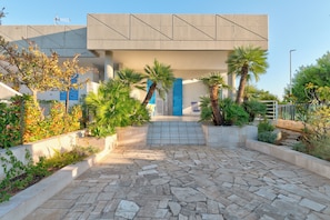A modern building with a concrete facade, blue shutters, and a stone-paved courtyard with palm trees.