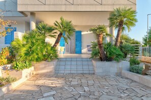 Un bâtiment moderne avec une entrée pavée de pierres, des volets bleus et des palmiers.