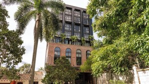 Um edifício moderno com base em tijolo e andares superiores escuros, rodeado por vegetação exuberante e palmeiras.