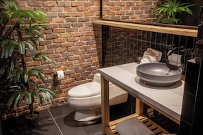 A bathroom with a brick wall, a toilet, a sink with a basin, and a shelf above the sink. There are two potted plants, one on the left and one on the right. - The Host Medellin  (Medellín)