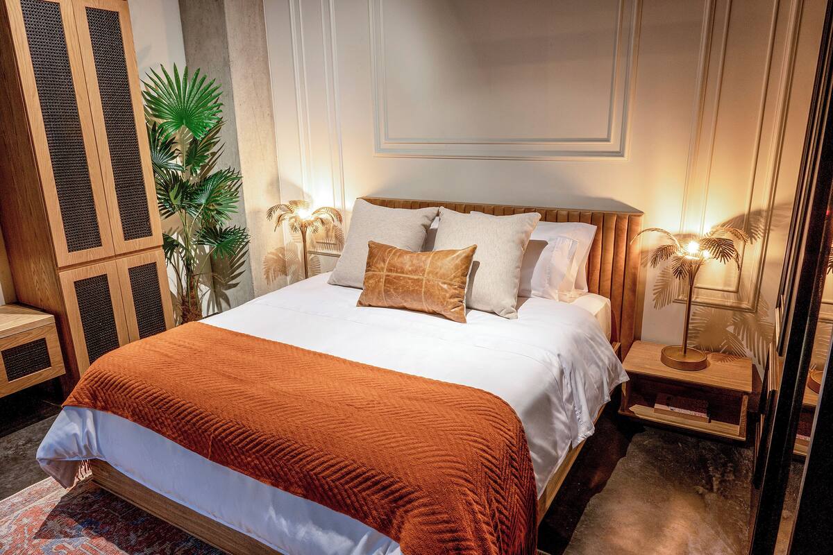 Een net opgemaakt bed met een wit-oranje sprei, twee kussens en een houten hoofdbord. Er staan twee nachtkastjes met lampen en een potplant naast het bed.
