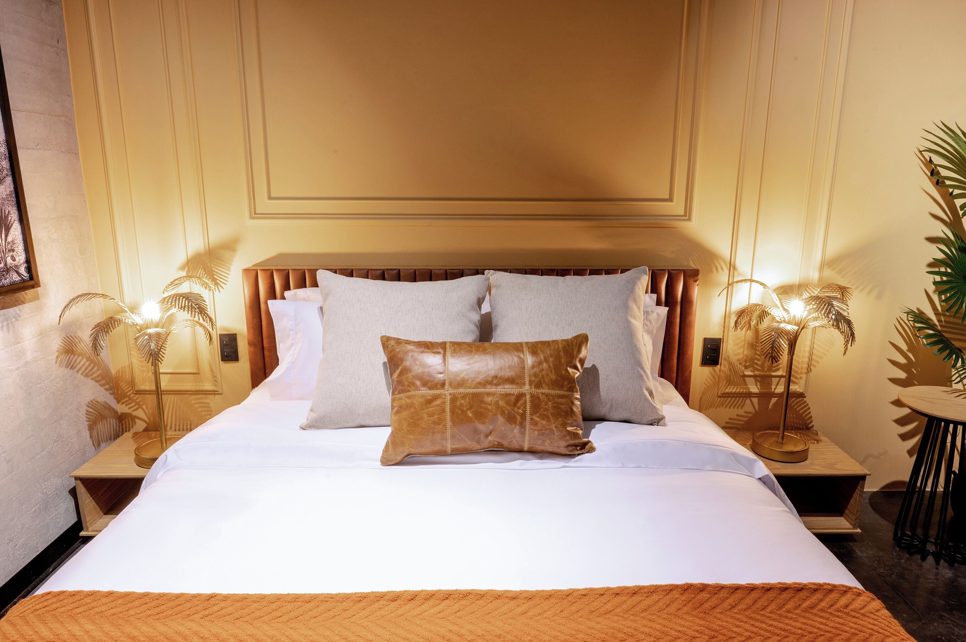 A neatly made bed with a brown leather pillow, white linens, and two matching pillows. There are two bedside tables with lamps, a potted plant, and a small round table with a chair.