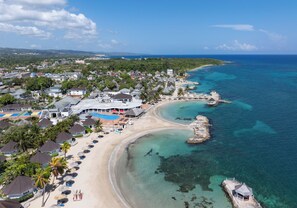 Vue aérienne d’une station balnéaire dotée d’un accès à la plage, d’eaux cristallines et de divers bâtiments, notamment un hôtel et des immeubles résidentiels.