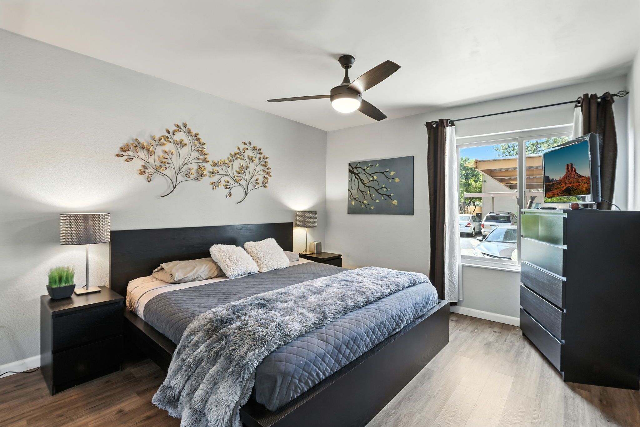 Un dormitorio moderno con cama, mesitas de noche, ventilador de techo, obras de arte y vistas al exterior.