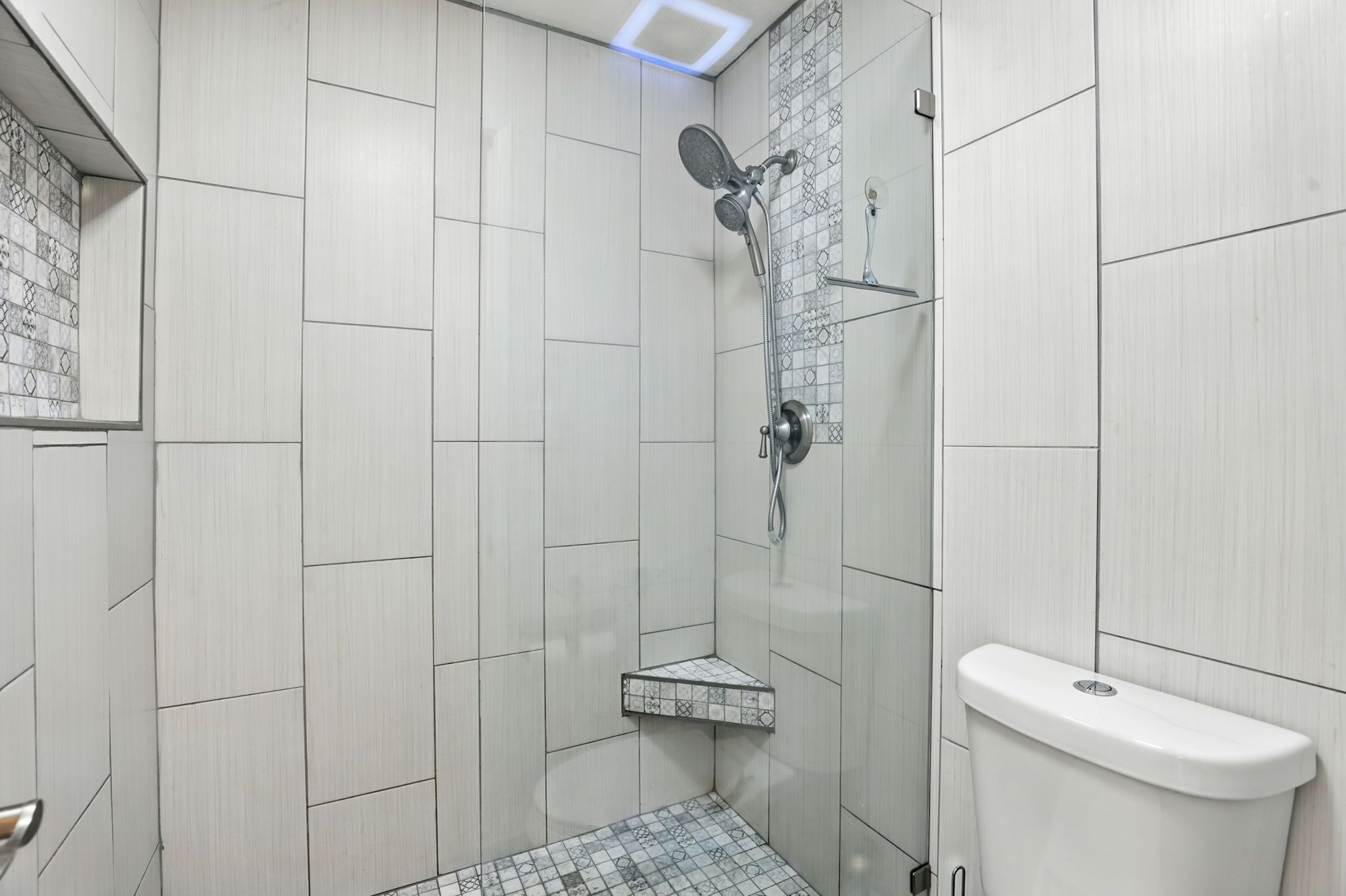 A modern bathroom with white tiles, a shower with a handheld showerhead, and a toilet.