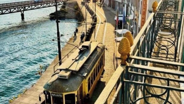 Eine Straßenbahn auf Gleisen entlang eines Flusses, im Hintergrund eine Brücke und Gebäude mit Balkonen.