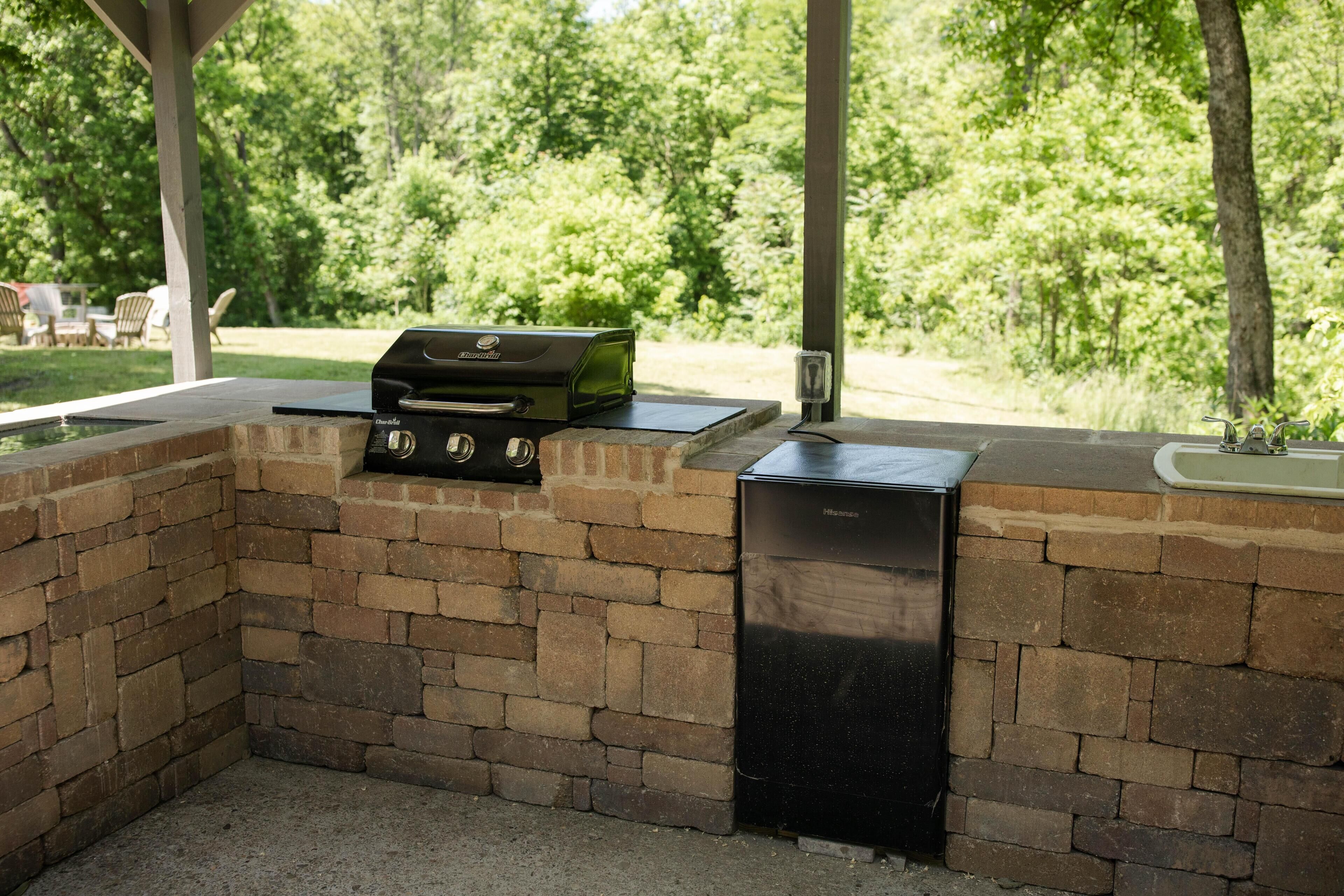 Cocina exterior con parrilla y lavavajillas, rodeada por un muro de ladrillo y vistas a un bosque verde y exuberante.