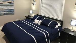 A bedroom with a navy blue bedspread, a nightstand with a lamp, a painting on the wall, and a window with blinds. - Algonac Harbour House (Algonac)