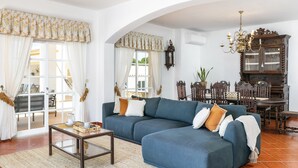 Ein Wohnzimmer mit einer Eckcouch, einem Couchtisch und einer Deckenleuchte. Im Hintergrund steht ein Esstisch mit Stühlen und ein Schrank.